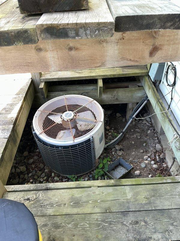 Responded to no cooling call on central air conditioning system over 15 years old. Upon arrival, located outdoor condensing unit installed beneath deck structure in recessed area. Performed diagnostic evaluation of system components and documented equipment condition with photos showing cylindrical condenser with copper fan guard, black vertical fins, refrigerant lines, and electrical disconnect box mounted on wooden framing.