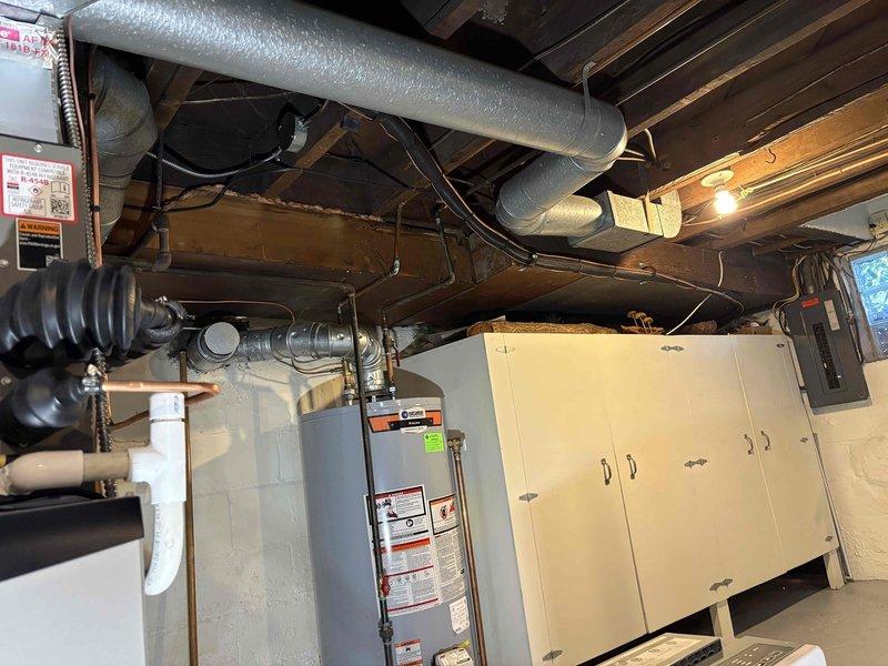Completed full HVAC system installation in basement utility area, including 70,000 BTU furnace with exposed control panel and circuit board section, 2-ton air conditioning unit with rear placement configuration, and humidifier integration. Installed white PVC venting system extending from furnace to ceiling with proper exhaust routing, connected silver metallic supply and return ductwork with vertical-to-horizontal elbow transitions, and established all electrical connections with black cabling routed along ceiling joists. Reconnected existing utilities and verified proper operation of all mechanical systems. All equipment positioned on concrete floor with adequate clearance for service access, permits obtained prior to installation, and system tested for proper airflow and functionality.