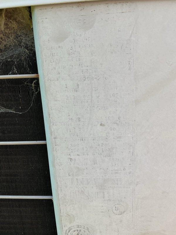Responded to service call for 20+ year old outdoor air conditioning unit with suspected refrigerant leak in coil. Upon arrival, performed visual inspection of condenser unit positioned against building exterior. Noted unit showing significant age-related wear with dirty condenser coils containing accumulated debris, cobwebs, and fibrous material between fins. Information plate on unit panel heavily weathered with faded specifications. Assessed system for refrigerant leak as reported, examining evaporator coil condition and checking for signs of refrigerant loss. Unit requires thorough coil cleaning to restore proper airflow and heat transfer efficiency before further diagnostics can determine extent of refrigerant system issues.