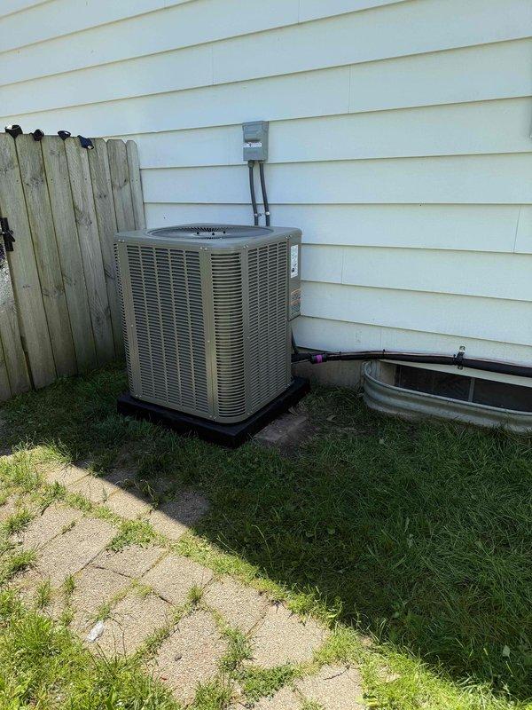 Completed full HVAC system installation including outdoor condensing unit and indoor furnace. Mounted beige residential air conditioning condenser on exterior wall with proper mounting pad and installed gray electrical disconnect box with appropriate conduit routing to unit. Positioned equipment against vinyl siding with adequate clearance for service access and airflow. In basement mechanical room, installed new gray furnace unit with complete metal ductwork routing and proper ventilation configuration. Connected all refrigerant lines between outdoor and indoor components. Verified electrical connections, tested system operation, and confirmed proper airflow through ductwork. System commissioned and operating within manufacturer specifications.