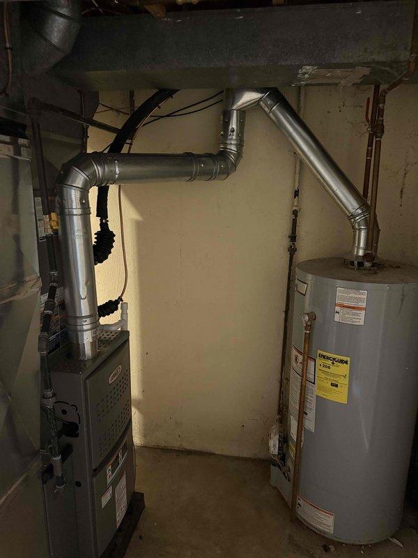 Completed full HVAC system installation including outdoor condensing unit and indoor furnace. Mounted beige residential air conditioning condenser on exterior wall with proper mounting pad and installed gray electrical disconnect box with appropriate conduit routing to unit. Positioned equipment against vinyl siding with adequate clearance for service access and airflow. In basement mechanical room, installed new gray furnace unit with complete metal ductwork routing and proper ventilation configuration. Connected all refrigerant lines between outdoor and indoor components. Verified electrical connections, tested system operation, and confirmed proper airflow through ductwork. System commissioned and operating within manufacturer specifications.