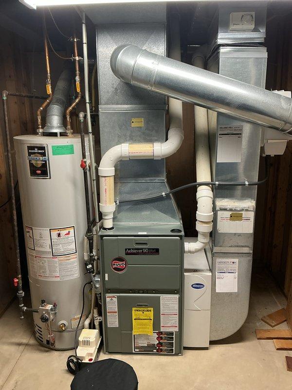 Completed comprehensive air conditioning maintenance on 19-year-old Amana condensing unit and Ruud Achiever 90 Plus furnace system under membership agreement. Performed full AC tune-up including inspection of outdoor condensing unit showing age-related weathering and rust staining on lower cabinet and grille sections, verified refrigerant levels, cleaned condenser coils, checked electrical connections and capacitor operation, and inspected fan motor assembly. Evaluated indoor air handler components, examined ductwork connections for proper sealing, verified condensate drain lines and PVC drainage system for proper flow, tested thermostat operation and system cycling. Confirmed system operational at conclusion of maintenance with all components functioning within normal parameters despite advanced age of equipment. Documented equipment condition with photographs showing outdoor unit on concrete pad with river rock landscaping and indoor mechanical room installation. System ready for coo