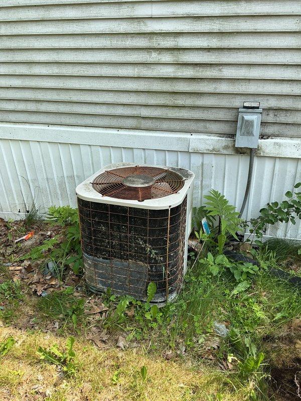 Responded to service call for 10-15 year old outdoor AC unit not blowing cold air. Upon arrival, found cylindrical condenser unit in severely deteriorated condition with extensive rust on protective cage, heavy dirt and grime accumulation, and dense vegetation growth obstructing airflow around base and sides. Cleared overgrown weeds, grass, and ferns from around unit perimeter to restore proper ventilation clearance. Cleaned exterior surfaces of condenser to remove accumulated debris, dirt, and biological growth that was restricting airflow and heat transfer efficiency. Assessed protective wire cage showing significant corrosion and oxidation throughout. Unit operational following cleaning and vegetation removal, with improved airflow restored to condenser.