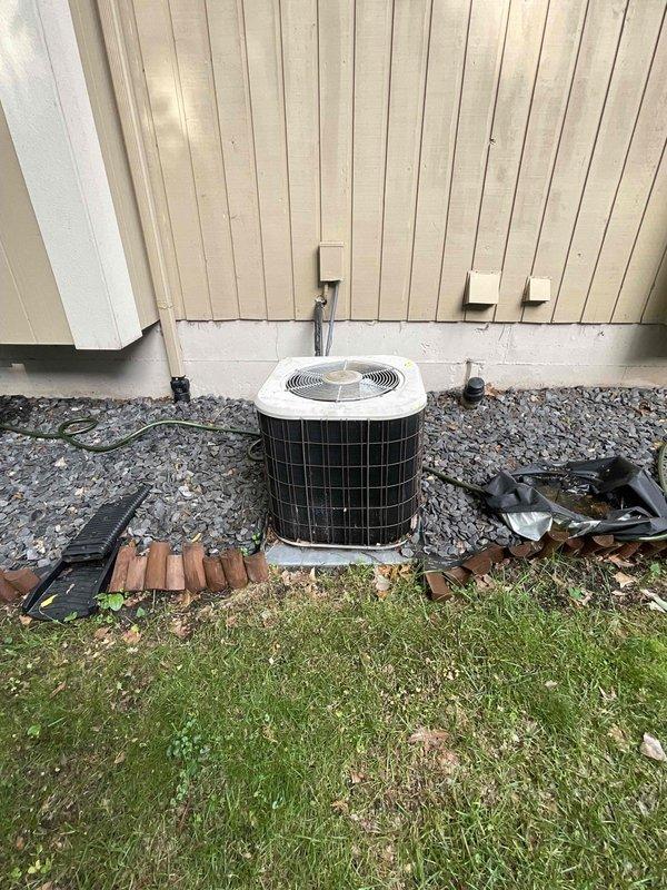 Conducted on-site assessment for complete HVAC system replacement estimate at approximately 1,900 sq ft single-family residence. Evaluated existing outdoor air conditioning condenser unit positioned on concrete pad with gravel surround against exterior wall, and basement-located gas furnace of unknown age. Documented current equipment configuration including electrical service, refrigerant line routing, and duct transitions. Photographed existing condenser installation showing white cabinet unit with protective top grille, electrical conduit and disconnect box mounting, and surrounding site conditions. Prepared comprehensive replacement quote for cost-effective system including new air conditioning unit and gas furnace, complete removal and disposal of existing equipment, installation of standard refrigerant line set, duct transition work, permit acquisition and inspection coordination, basic thermostat provision if required, and full warranty coverage details.