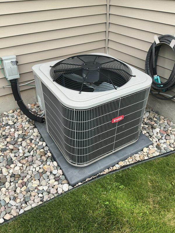 Completed annual cooling maintenance on residential HVAC system. Inspected outdoor condensing unit with white casing and confirmed proper operation of fan motor and compressor. Checked refrigerant lines running along exterior wall for secure connections and proper insulation. Verified electrical disconnect box functionality and examined all external components for wear or damage. Inspected indoor Bryant furnace unit in basement mechanical room, including air handler components and PVC condensate drainage system. Documented equipment condition through photographs showing well-maintained installation with proper clearances and landscaping integration around outdoor unit on protective pad with decorative stone border.