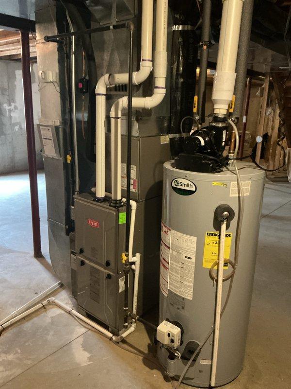 Completed annual cooling maintenance on residential HVAC system. Inspected outdoor condensing unit with white casing and confirmed proper operation of fan motor and compressor. Checked refrigerant lines running along exterior wall for secure connections and proper insulation. Verified electrical disconnect box functionality and examined all external components for wear or damage. Inspected indoor Bryant furnace unit in basement mechanical room, including air handler components and PVC condensate drainage system. Documented equipment condition through photographs showing well-maintained installation with proper clearances and landscaping integration around outdoor unit on protective pad with decorative stone border.