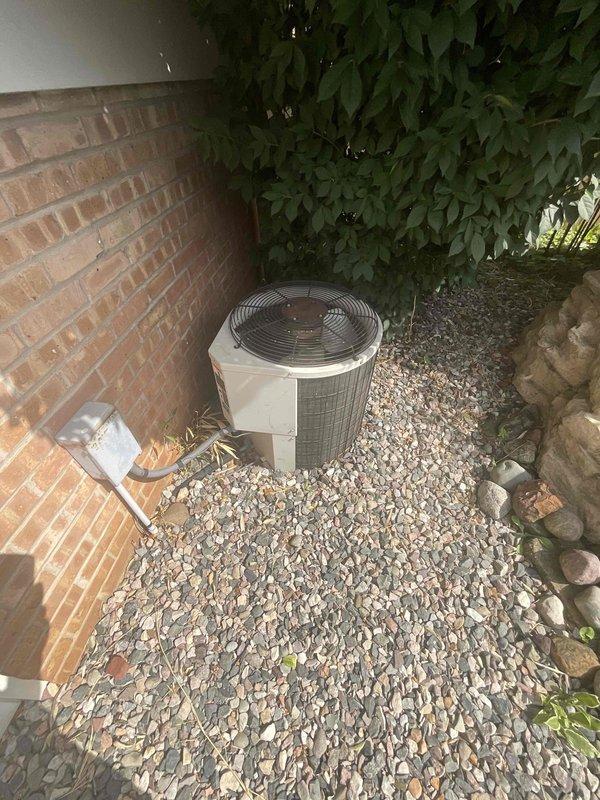 Conducted estimate for replacement of 40-year-old outdoor air conditioning condenser unit currently positioned on gravel bed adjacent to brick wall with limited side yard access. Assessed existing cylindrical unit with beige housing and top-mounted fan assembly, noting age-related equipment obsolescence and customer's request for expedited replacement. Documented site conditions including narrow installation space between structure and landscaping, gravel base pad, and electrical service connection at brick wall for replacement planning and equipment sizing recommendations.