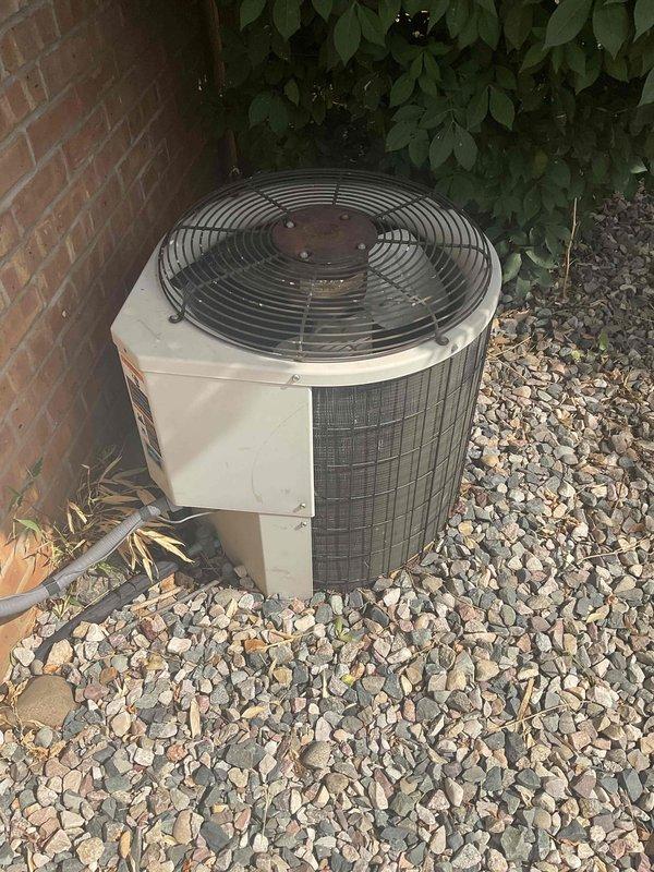 Conducted estimate for replacement of 40-year-old outdoor air conditioning condenser unit currently positioned on gravel bed adjacent to brick wall with limited side yard access. Assessed existing cylindrical unit with beige housing and top-mounted fan assembly, noting age-related equipment obsolescence and customer's request for expedited replacement. Documented site conditions including narrow installation space between structure and landscaping, gravel base pad, and electrical service connection at brick wall for replacement planning and equipment sizing recommendations.