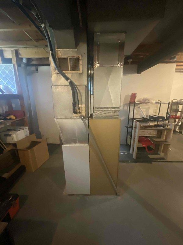 Completed HVAC system replacement estimate for duplex unit, documenting existing furnace and air handler equipment in basement mechanical room. Photographed current system configuration including gray metal furnace unit with horizontal grille vent, cream-colored air handler with metallic exhaust piping, black flexible ductwork routing, and electrical panel connections to assess replacement requirements and system specifications.