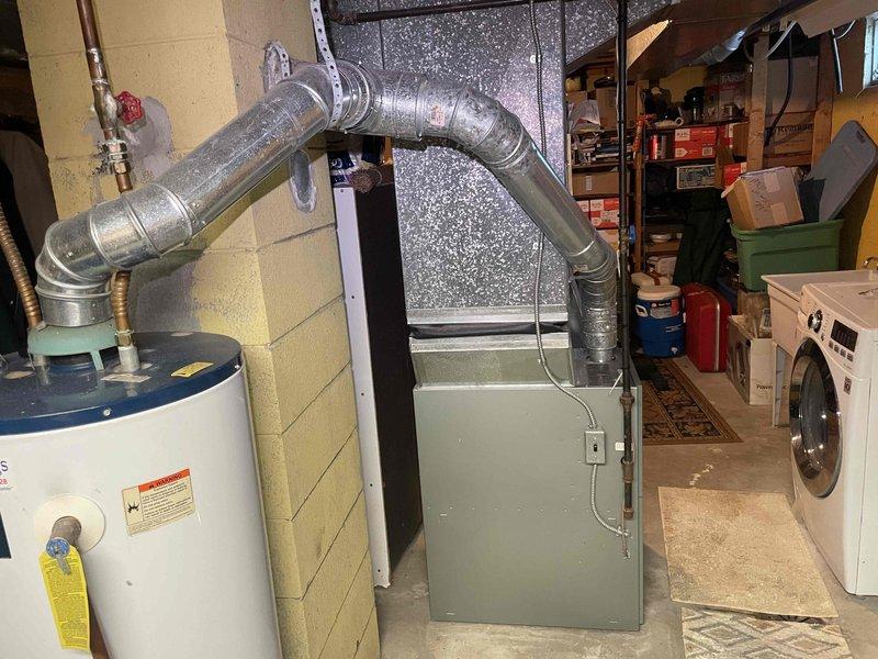 Conducted site assessment for central air conditioning installation in a 1970s home currently equipped with a Ruud Silhouette II gas furnace but no existing AC system. Evaluated basement mechanical room layout, existing ductwork configuration, and available space for condenser and air handler placement. Photographed current HVAC setup including furnace location, flexible ductwork routing, water heater proximity, and utility room constraints to develop comprehensive whole-house cooling solution estimate.