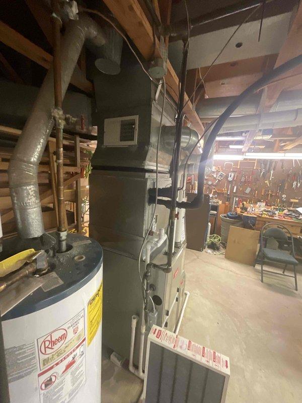 Responded to customer request for air conditioning replacement due to water leaking under furnace during cooling operation. Inspected 21-year-old HVAC system located in basement alongside Rheem water heater, noting aged furnace unit with metal ductwork extending through ceiling joists. Documented existing equipment configuration including electrical panel with circuit breakers and PVC plumbing runs for estimate preparation. Provided comprehensive replacement options for complete system upgrade to resolve water leakage issue and improve efficiency.