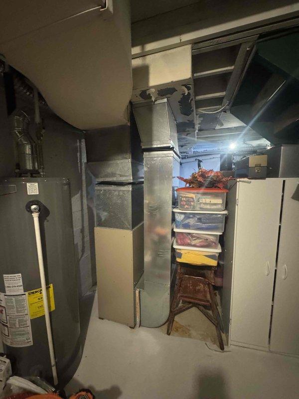 Conducted comprehensive site assessment for HVAC system replacement in residential basement utility area. Evaluated existing furnace unit, ductwork configuration, and available clearances for new equipment installation. Documented current system layout including water heater proximity, overhead ductwork routing, and structural considerations with exposed ceiling joists. Photographed existing beige furnace with connected metal supply ductwork and identified spatial constraints due to adjacent storage shelving and utility equipment. Assessed ceiling damage areas that may require attention during installation process. Provided detailed replacement estimate including equipment specifications, labor requirements, and installation scope for complete HVAC system upgrade.