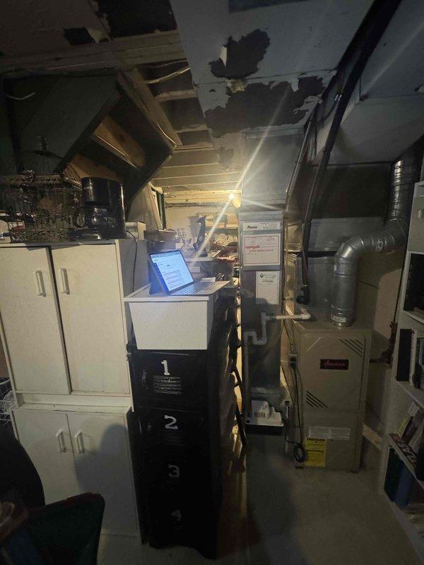 Conducted comprehensive site assessment for HVAC system replacement in residential basement utility area. Evaluated existing furnace unit, ductwork configuration, and available clearances for new equipment installation. Documented current system layout including water heater proximity, overhead ductwork routing, and structural considerations with exposed ceiling joists. Photographed existing beige furnace with connected metal supply ductwork and identified spatial constraints due to adjacent storage shelving and utility equipment. Assessed ceiling damage areas that may require attention during installation process. Provided detailed replacement estimate including equipment specifications, labor requirements, and installation scope for complete HVAC system upgrade.