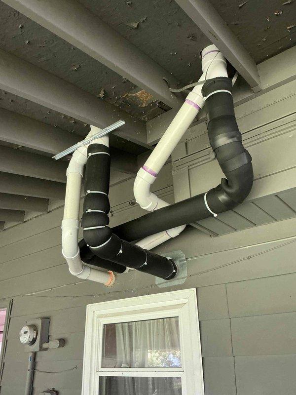 Completed callback service to reconfigure tankless water heater venting system by downsizing from 4" to 3" PVC and installing concentric vent piping through the overhang. Rerouted one of the concentric vents to maintain proper spacing from the adjacent vent line and corrected an intake section that had developed a trap condition. The new 3" concentric vent configuration now properly penetrates the covered porch area with adequate clearance and proper pitch to prevent condensate accumulation.