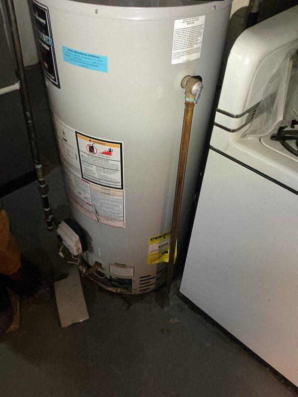 Responded to water heater leak reported at base of unit in 53-year-old residence. Upon inspection, confirmed active leak contained to area directly beneath tanked water heater in utility room. Observed visible water staining and discoloration on concrete floor surrounding base of unit. Examined copper supply piping, brass fittings, and gas shutoff valve assembly with yellow handle at top connections. Assessed overall condition of aging water heater installation, noting oxidation and patina on copper pipes consistent with original 1973 home construction. Documented equipment condition and leak source with photographs showing full unit, piping configuration, and affected floor area. Leak appears to originate from tank itself rather than supply line connections or pressure relief valve, indicating probable tank failure requiring replacement.
