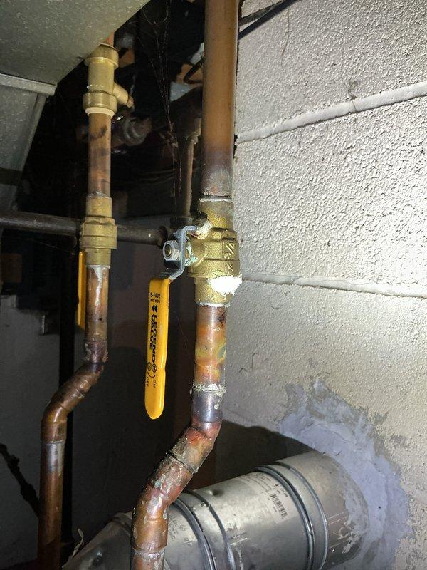 Responded to water heater leak reported at base of unit in 53-year-old residence. Upon inspection, confirmed active leak contained to area directly beneath tanked water heater in utility room. Observed visible water staining and discoloration on concrete floor surrounding base of unit. Examined copper supply piping, brass fittings, and gas shutoff valve assembly with yellow handle at top connections. Assessed overall condition of aging water heater installation, noting oxidation and patina on copper pipes consistent with original 1973 home construction. Documented equipment condition and leak source with photographs showing full unit, piping configuration, and affected floor area. Leak appears to originate from tank itself rather than supply line connections or pressure relief valve, indicating probable tank failure requiring replacement.