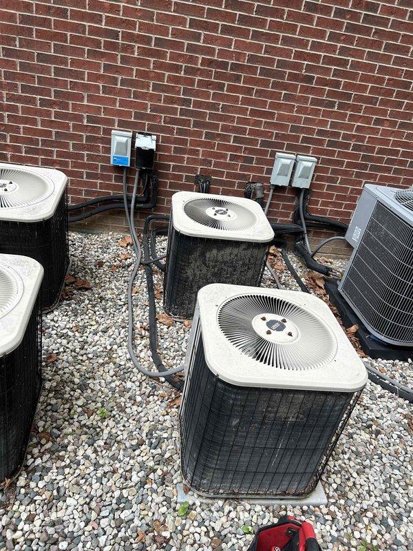 # HVAC Service Completed

Responded to no-cooling complaint on central air conditioning system at multi-unit property with five outdoor condenser units installed on gravel pad against brick exterior wall. Upon arrival, performed visual inspection of all condensing units, noting accumulated debris including dead leaves around equipment and some weathering on unit housings. 

Verified electrical connections at wall-mounted disconnect boxes and traced refrigerant line sets and conduit routing up brick wall. Assessed operational status of affected unit among the bank of four white-grilled square condensers and one silver/gray unit. Checked fan motor operation, compressor function, and electrical supply to identify cause of cooling failure. 

Cleared debris from around condenser units to ensure proper airflow and documented existing conditions of outdoor equipment installation. System evaluated for refrigerant levels, electrical issues, and mechanical component failure. Photographic documen