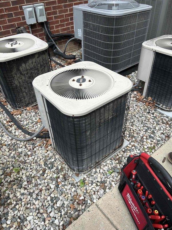 # HVAC Service Completed

Responded to no-cooling complaint on central air conditioning system at multi-unit property with five outdoor condenser units installed on gravel pad against brick exterior wall. Upon arrival, performed visual inspection of all condensing units, noting accumulated debris including dead leaves around equipment and some weathering on unit housings. 

Verified electrical connections at wall-mounted disconnect boxes and traced refrigerant line sets and conduit routing up brick wall. Assessed operational status of affected unit among the bank of four white-grilled square condensers and one silver/gray unit. Checked fan motor operation, compressor function, and electrical supply to identify cause of cooling failure. 

Cleared debris from around condenser units to ensure proper airflow and documented existing conditions of outdoor equipment installation. System evaluated for refrigerant levels, electrical issues, and mechanical component failure. Photographic documen