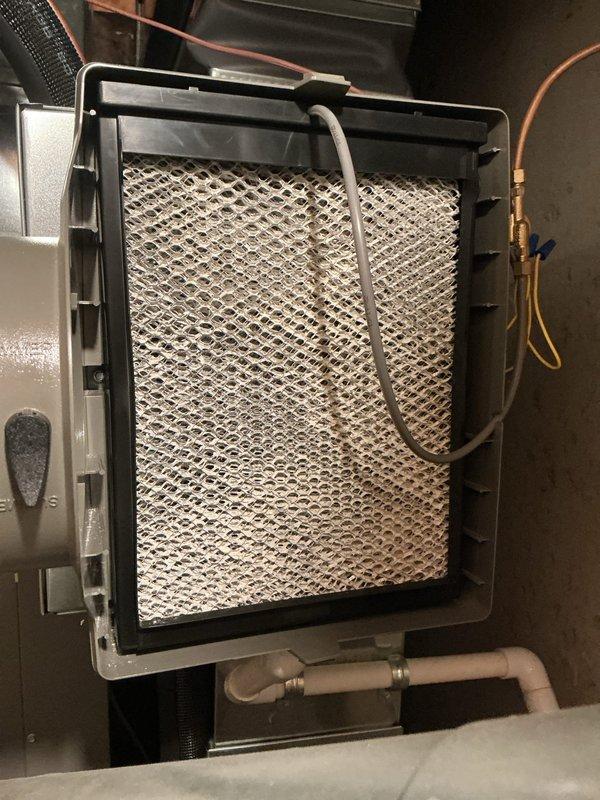 Completed scheduled maintenance on HVAC system including replacement of humidifier pad and air filter. The humidifier pad showed mineral deposits and discoloration from normal use and was replaced with new pad in the black plastic housing frame. Verified proper water distribution through the flexible tubing and confirmed system operation. Thermostat displaying normal cooling operation at 76° setpoint with outdoor temperature at 84°.