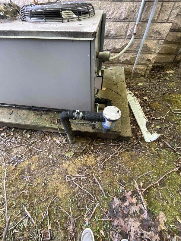 Provided estimate for replacement of non-operational residential air conditioning system serving basement area of cemetery mausoleum. Existing outdoor condensing unit shows significant age and weathering, with moss and algae growth on concrete pad and wooden platform, indicating prolonged exposure to moisture and lack of recent maintenance. Assessed equipment location, existing refrigerant line routing, electrical disconnect, and site conditions to determine appropriate replacement specifications and installation requirements for new HVAC system.