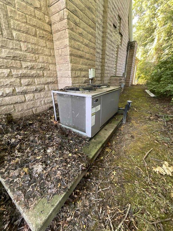 Provided estimate for replacement of non-operational residential air conditioning system serving basement area of cemetery mausoleum. Existing outdoor condensing unit shows significant age and weathering, with moss and algae growth on concrete pad and wooden platform, indicating prolonged exposure to moisture and lack of recent maintenance. Assessed equipment location, existing refrigerant line routing, electrical disconnect, and site conditions to determine appropriate replacement specifications and installation requirements for new HVAC system.