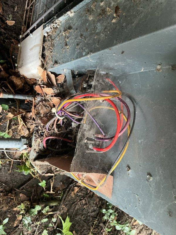 Responded to service call for outdoor AC condenser unit running loudly but still cooling effectively. Upon inspection, found significant debris accumulation around unit base including leaves, twigs, and vegetation growth impeding proper airflow. Observed exposed electrical wiring emerging from damaged section of base housing, creating potential safety hazard. Cleared organic debris from around condenser unit perimeter and verified fan operation and airflow through coil sections. Condenser coils showed weathering and dirt buildup on exterior surfaces. Advised customer that exposed wiring requires proper sealing and that debris accumulation likely contributing to excessive noise levels, though compressor and fan assembly continuing to function and maintain adequate cooling capacity.