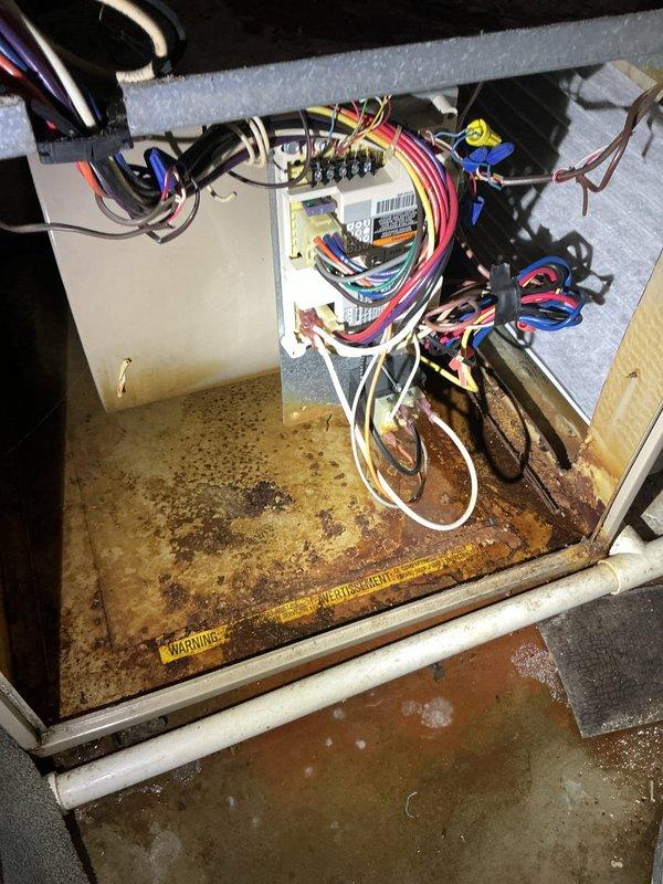Responded to service call for leaking HVAC system on 20-year-old unit still providing heating and cooling operation. Upon inspection, discovered significant water damage and corrosion in the drain pan area, with visible rust deterioration and moisture accumulation at the base of the unit. Photographed the interior compartment showing corroded drain pan with brownish-orange oxidation, standing water residue, and water staining throughout the affected area. Documented gas valve assembly, copper piping connections, electrical control panel with wiring harness, and overall internal components. Unit showed signs of age-related wear including dust accumulation and general weathering. The heavily corroded drain pan indicates ongoing moisture issues that require attention to prevent further deterioration and potential system failure.