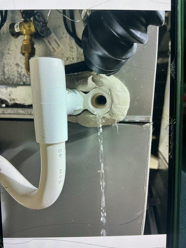 Responded to service call for non-functioning forced air furnace with reported water leakage down sides of unit. Upon inspection, identified condensate drainage issue affecting four-year-old system located in basement. Documented active water dripping from drain fitting above condensate trap assembly, with visible water stream bypassing proper drainage path through white PVC P-trap configuration. Examined HVAC unit labels confirming system specifications and proper refrigerant warnings. Inspected condensate removal system including PVC piping, copper line connections, and drainage components. Photographed plumbing infrastructure showing compromised drain fitting causing water intrusion. Assessed P-trap assembly, standpipe configuration, and associated drainage hardware to determine source of moisture problem affecting furnace operation.
