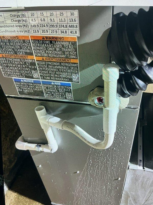 Responded to service call for non-functioning forced air furnace with reported water leakage down sides of unit. Upon inspection, identified condensate drainage issue affecting four-year-old system located in basement. Documented active water dripping from drain fitting above condensate trap assembly, with visible water stream bypassing proper drainage path through white PVC P-trap configuration. Examined HVAC unit labels confirming system specifications and proper refrigerant warnings. Inspected condensate removal system including PVC piping, copper line connections, and drainage components. Photographed plumbing infrastructure showing compromised drain fitting causing water intrusion. Assessed P-trap assembly, standpipe configuration, and associated drainage hardware to determine source of moisture problem affecting furnace operation.