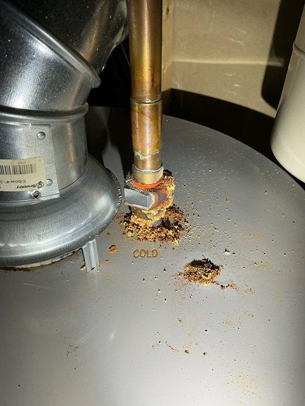 Responded to service call for inspection of traditional gas-fired water heater with reported corrosion on cold water supply line. Upon arrival, conducted thorough visual assessment of water heater and associated plumbing connections. Confirmed presence of moderate surface corrosion and rust deposits at the cold water inlet pipe connection point on top of the unit. Corrosion exhibited characteristic orange-brown oxidation with granular, crusty texture and material flaking around the copper pipe junction. Inspected all threaded brass fittings, ball valve assembly, and pipe connections for integrity and potential leak points. Verified no active water leaks, moisture accumulation, or dripping present at time of inspection. Examined PTFE tape application on threaded connections and confirmed proper sealing. Assessed overall condition of copper supply piping, noting age-related patina and surface discoloration consistent with normal wear. Documented corrosion location and extent through phot
