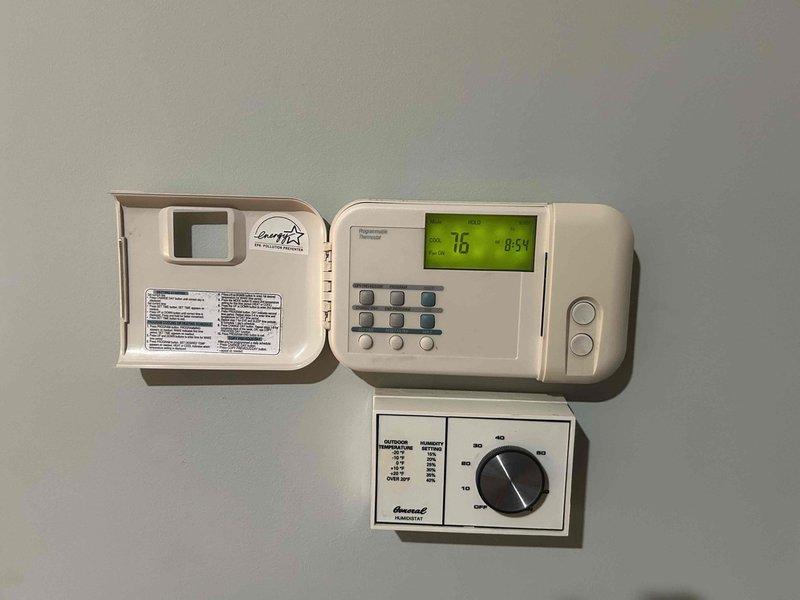 Responded to service call for 1997 A/C system in basement location reporting unusual humming noise and inadequate cooling performance. Upon inspection, accessed furnace cabinet to examine blower motor assembly, electrical connections, and control components. Evaluated thermostat operation and system controls, noting presence of older analog and digital programmable thermostats. Documented equipment condition through photography showing internal components including blower motor, gas valve assemblies, wiring, and control boards, as well as thermostat configuration displaying 76-degree reading at time of service.