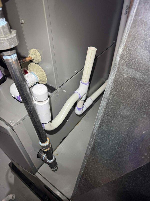 Responded to third callback for furnace water leak and noisy blower motor on 2.5-year-old forced air system. Inspected condensate drain system and PVC drainage lines with purple-primed connections at air handler cabinet. Verified proper pitch and drainage flow through white PVC piping system, checked insulated refrigerant lines and copper tubing connections for condensation issues. Documented current installation configuration showing drain lines, insulated suction line with foam wrap, and equipment connections. System operational at time of inspection with heating function confirmed.