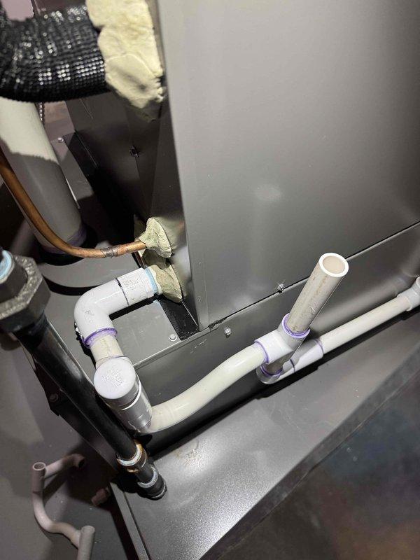 Responded to third callback for furnace water leak and noisy blower motor on 2.5-year-old forced air system. Inspected condensate drain system and PVC drainage lines with purple-primed connections at air handler cabinet. Verified proper pitch and drainage flow through white PVC piping system, checked insulated refrigerant lines and copper tubing connections for condensation issues. Documented current installation configuration showing drain lines, insulated suction line with foam wrap, and equipment connections. System operational at time of inspection with heating function confirmed.