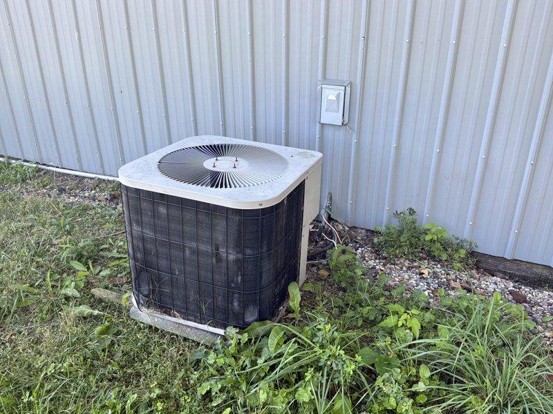 Provided estimate for complete HVAC system conversion from non-functional boiler heating to forced air furnace and air conditioning system. Existing outdoor condenser unit shows significant age and weathering with overgrown vegetation, while interior office space currently utilizes baseboard heating requiring full ductwork installation and equipment replacement for modernized climate control.