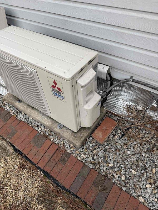 Conducted sales consultation for potential mini-split installation, with Aaron as tech lead. Documented existing Mitsubishi outdoor condensing unit mounted on concrete pad with appropriate stone drainage bed and brick paver border. System appears to be a standard residential split configuration with connections running to the interior of the home.
