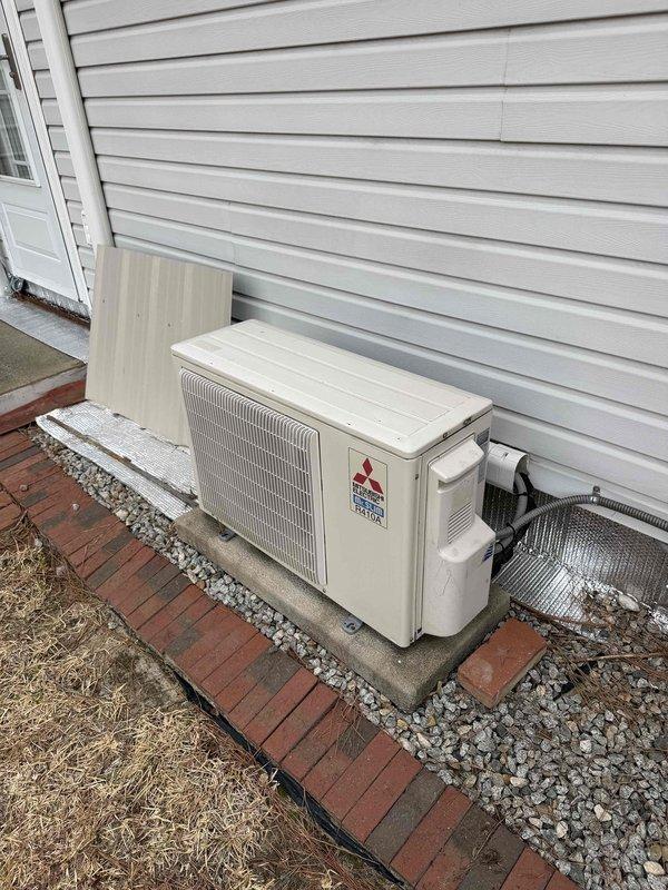 Conducted sales consultation for potential mini-split installation, with Aaron as tech lead. Documented existing Mitsubishi outdoor condensing unit mounted on concrete pad with appropriate stone drainage bed and brick paver border. System appears to be a standard residential split configuration with connections running to the interior of the home.