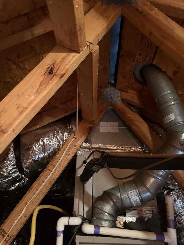 Conducted inspection of downstairs furnace reported with crack and non-operational status. Customer requested conversion from gas to electric heating system. Visual assessment of attic/crawl space revealed existing ductwork infrastructure with metal ventilation system, PVC plumbing, and electrical components integrated within wooden structural framework. Photos documented current installation conditions to support system replacement planning.