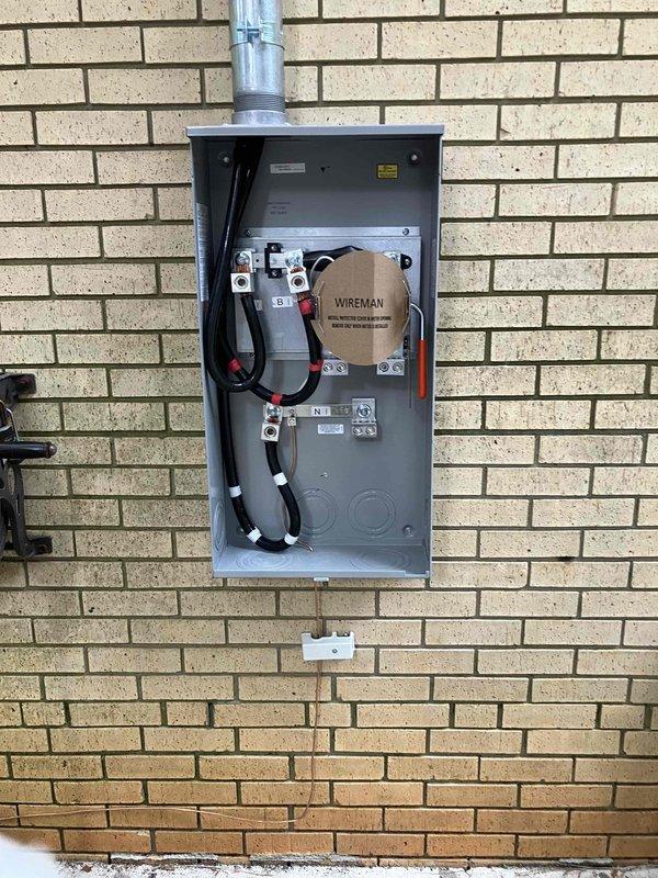 Completed return visit to verify meter base installation and document current status of service entrance. Meter enclosure previously installed on exterior brick wall with service entrance conductors terminated at terminal blocks, but final connections remain pending utility company engineering completion. Photographed installed meter base showing proper mounting, conduit routing to overhead service, and internal wiring configuration for documentation and coordination with utility provider.