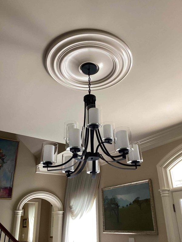 Installed customer-supplied chandelier with black metal frame and multiple glass shades in residential foyer with 10-foot ceiling. Mounted decorative white ceiling medallion with concentric circular pattern to ceiling junction box, secured chandelier chain and canopy, made electrical connections, and tested all lamp sockets for proper operation.