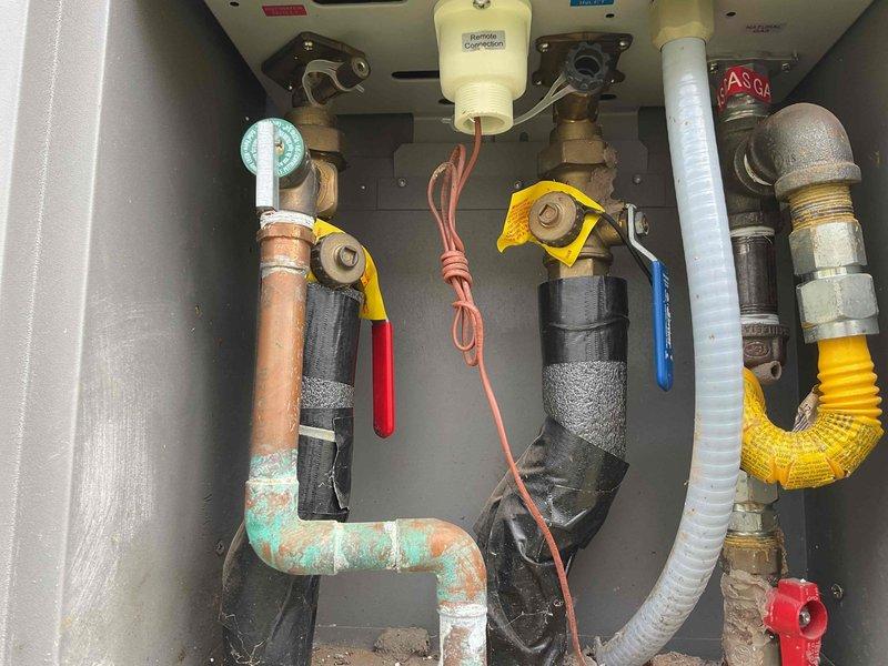 Responded to no hot water complaint on 7-year-old Rheem tankless gas water heater installed in exterior alcove. Upon arrival, performed diagnostic evaluation and found unit displaying error code indicating ignition failure. Inspected gas supply line, verified proper gas pressure at yellow corrugated flex line connection, and confirmed gas valve operation. Tested flow sensor functionality and checked water inlet filtration for blockages. Examined igniter assembly and flame rod for carbon buildup and proper positioning. Verified venting system integrity and checked for obstructions in intake and exhaust pathways. Assessed electrical connections, tested voltage supply, and confirmed proper grounding. Observed visible corrosion on copper water supply piping with green patina formation at elbow joints, noted insulation deterioration on multiple pipe sections. Documented pressure gauge readings and valve positions throughout plumbing connections within utility cabinet. Given unit age and dia
