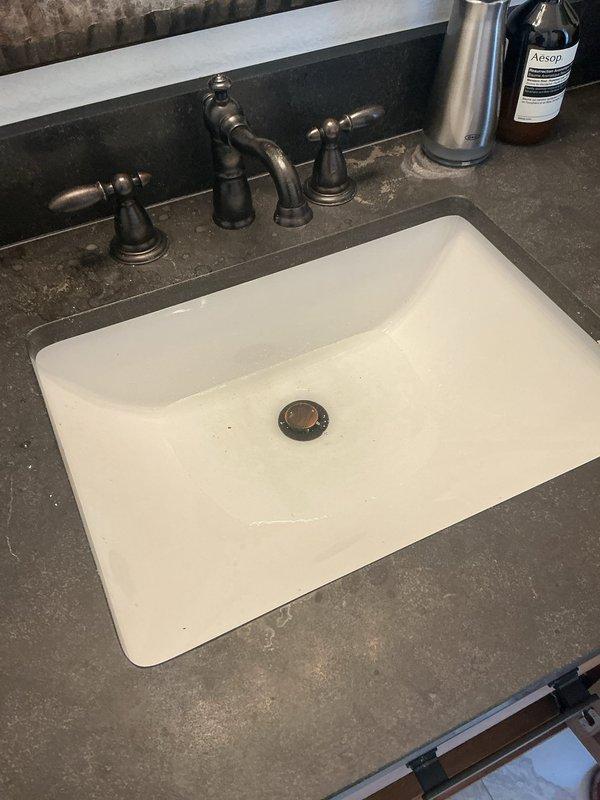 Responded to clogged bathroom sink in 1992 home. Upon inspection, found undermount white rectangular basin with bronze pop-up drain assembly and three-handle faucet fixture experiencing drainage issues. Accessed plumbing beneath sink and cleared obstruction from P-trap assembly. Verified proper drainage flow through tailpiece and trap configuration. Tested sink operation and confirmed water drains freely with no backup or slow drainage remaining.