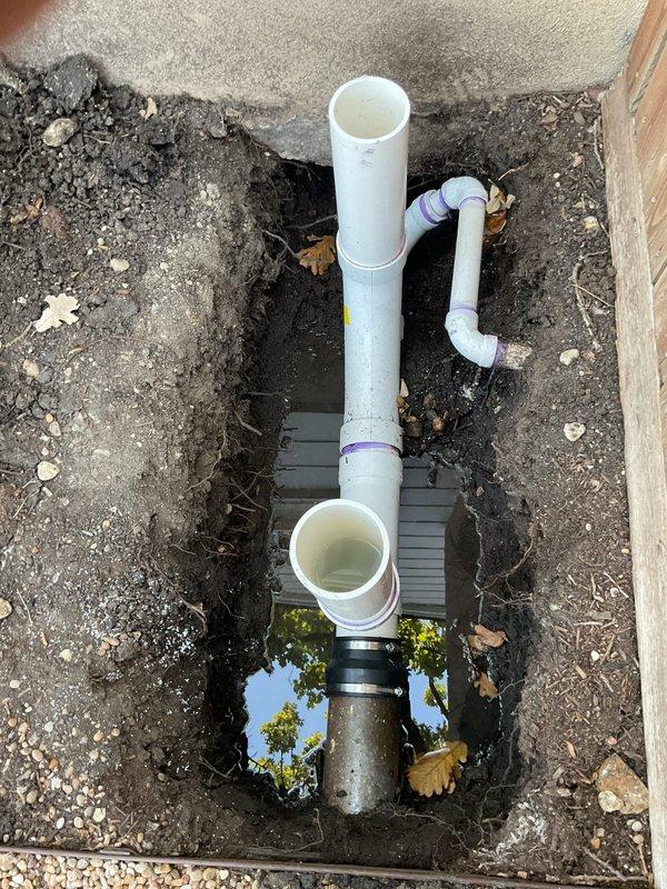Responded to emergency drainage service call at 1959 residence where main sewer line work by another contractor (SALT) resulted in non-functioning toilets and flooding from exposed sewer drains. Accessed main line cleanout located in crawlspace beneath home as homeowner was not present. Inspected underground drainage system and documented PVC pipe configuration including vertical stacks, horizontal runs, and connection points with brass fittings at exterior access points. Found standing water accumulation in excavated utility pit indicating active drainage issues. Assessed exterior plumbing installation where building foundation meets ground level, including cleanout access valve and associated piping. Photographed existing conditions showing water pooling, exposed soil around pipe penetrations, and current state of drain line assembly for documentation and coordination with ongoing contractor work.