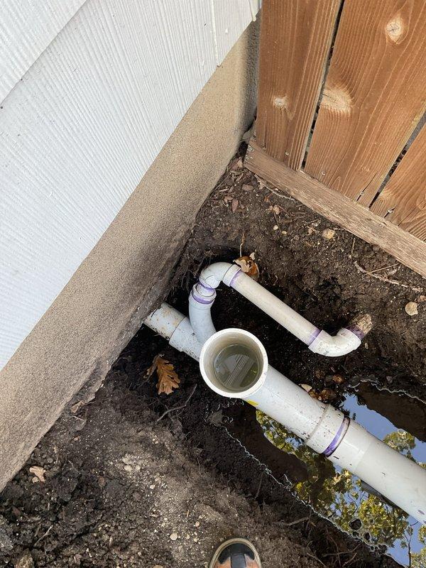 Responded to emergency drainage service call at 1959 residence where main sewer line work by another contractor (SALT) resulted in non-functioning toilets and flooding from exposed sewer drains. Accessed main line cleanout located in crawlspace beneath home as homeowner was not present. Inspected underground drainage system and documented PVC pipe configuration including vertical stacks, horizontal runs, and connection points with brass fittings at exterior access points. Found standing water accumulation in excavated utility pit indicating active drainage issues. Assessed exterior plumbing installation where building foundation meets ground level, including cleanout access valve and associated piping. Photographed existing conditions showing water pooling, exposed soil around pipe penetrations, and current state of drain line assembly for documentation and coordination with ongoing contractor work.