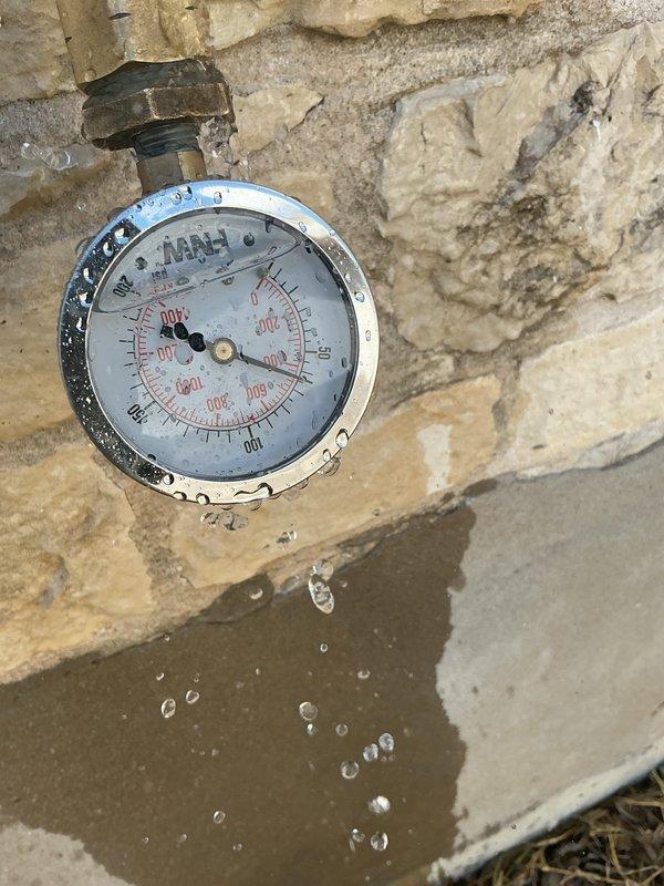 Responded to an active water leak at a garage pressure gauge assembly. Upon arrival, found water actively dripping from a brass threaded fitting at the wall connection point where a pressure gauge was mounted. The gauge displayed significant weathering and corrosion on the chrome finish, and a puddle had formed on the concrete floor beneath the fixture. 

Inspected the CPVC plumbing system configuration on the adjacent wall, which included vertical and horizontal pipe sections connected by elbow joints with black rubber coupling connectors. Given the home's age and the condition of the pressure gauge assembly, determined that the leak originated from a failed seal at the threaded connection between the brass fitting and the wall-mounted pipe. 

Shut off water supply to the affected line and removed the deteriorated pressure gauge. Cleaned the threaded brass fitting and inspected for damage or corrosion that could prevent proper resealing. Applied appropriate thread sealant and reinstal