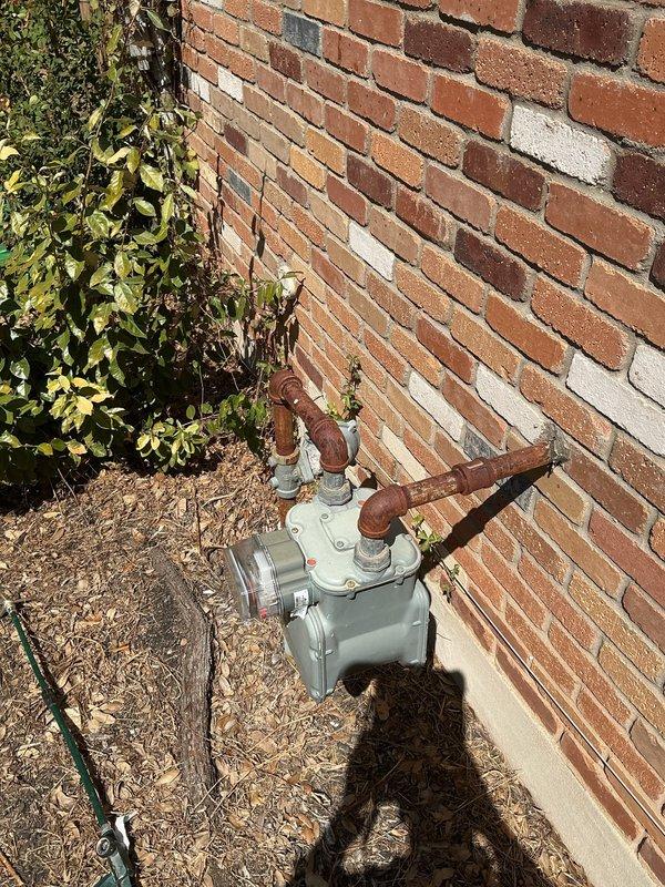 Responded to customer concern regarding humming and vibrating natural gas meter. Upon arrival, inspected exterior gas meter installation mounted on brick foundation. Examined meter body, regulator assembly, and associated copper gas piping for loose connections, proper mounting, and structural integrity. Verified all pipe connections, elbows, and shut-off valve were secure and properly supported. Assessed meter mounting bracket and foundation stability. Documented existing piping configuration showing weathered copper lines with oxidation and multiple connection points. No immediate safety hazards or gas leaks detected during visual inspection.