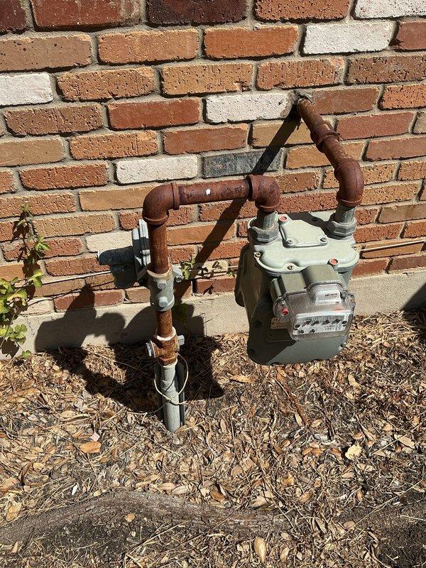 Responded to customer concern regarding humming and vibrating natural gas meter. Upon arrival, inspected exterior gas meter installation mounted on brick foundation. Examined meter body, regulator assembly, and associated copper gas piping for loose connections, proper mounting, and structural integrity. Verified all pipe connections, elbows, and shut-off valve were secure and properly supported. Assessed meter mounting bracket and foundation stability. Documented existing piping configuration showing weathered copper lines with oxidation and multiple connection points. No immediate safety hazards or gas leaks detected during visual inspection.