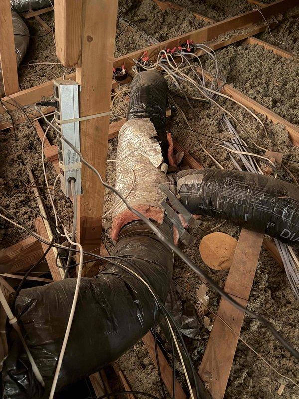 Performed an on-site evaluation of the attic duct system to provide a replacement estimate for the existing flexible ductwork. Inspection found flex duct jackets torn open at a Y-branch junction with exposed fiberglass insulation and foil liner, temporary tape repairs, crinkled and loosely bunched vapor-barrier wrap at plenum connections, and weathered, worn insulation on the rectangular plenum. Documented conditions with photos, noting loose wiring and debris throughout the attic, and prepared the findings for a duct replacement proposal.