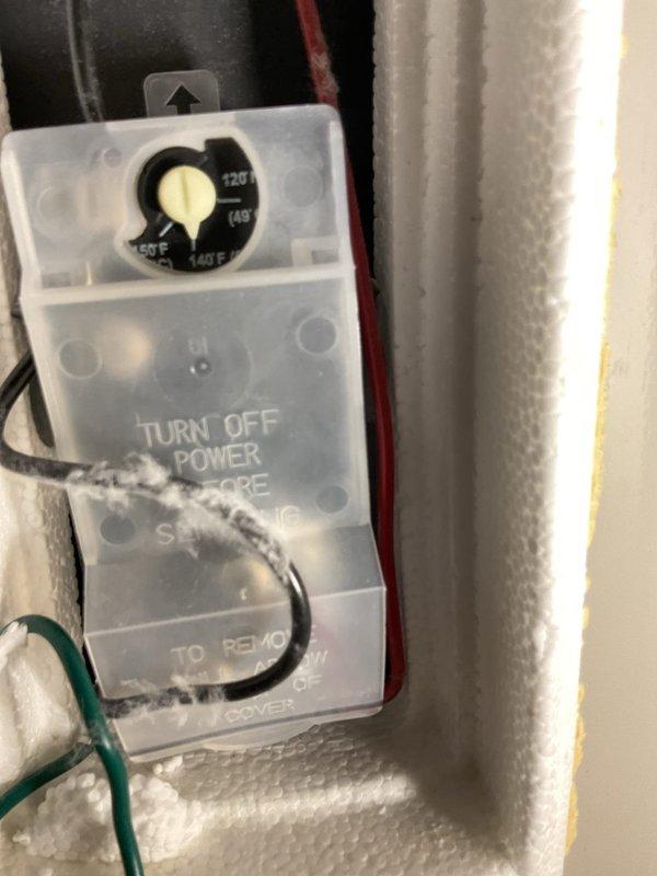 Evaluated an approximately 19-year-old electric tank water heater in the garage that was not producing hot water. Inspected the thermostat assembly and found the thermostat cover with a lint-coated wire and surrounding foam insulation intact, with no evidence of frost, cracks, or rust at the control compartment. Also inspected the thermal expansion tank mounted above the unit and found corrosion and paint loss on the tank body, indicating deterioration of the tank and the unit's advanced age and end of service life.