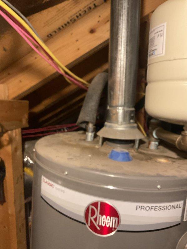 Responded to a reported leak in the bathroom walls and inspected the plumbing system, including the attic-mounted gas tank water heater with its flue, draft hood, insulated supply piping, and expansion tank connection, along with the exterior PVC sleeve and buried valve fitting, noting visible rust staining and soil and debris accumulation at the exterior fitting. Findings were documented with photographs for further evaluation and recommended repair planning.