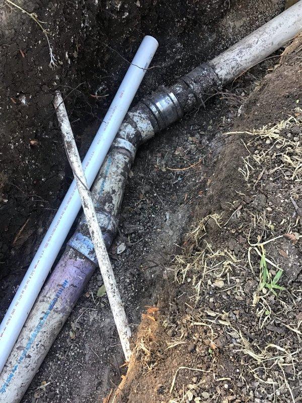 Excavated a trench to expose the underground drain line and completed the drain tunnel work, including installation of PVC drainage piping with a 90-degree elbow fitting and a rubber-banded coupling transition to larger sewer-style pipe. Verified pipe alignment and fitting connections within the trench, with no visible cracking or damage observed on the exposed piping.